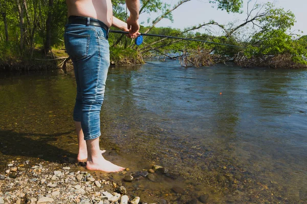 一个穿着牛仔裤的渔夫站在山河岸边的水里 他拿着一根钓竿 在水里漂浮着 — 图库照片