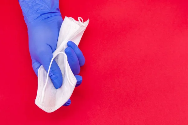 Tıbbi maske, covid-19, mavi tek kullanımlık eldivenli bir el kırmızı arka planda beyaz bir maske tutuyor, Coronavirus 'un sonu kavramı..