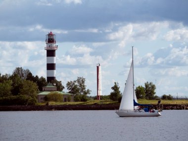 Yat nehir boyunca gider, güneşli bir gün ve anlamlı bulutlar. Arka planda deniz feneri olan bir kıyı şeridi.