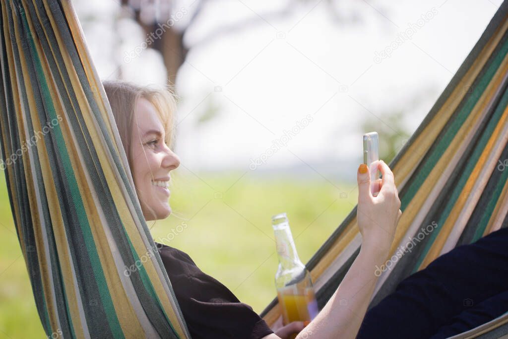 A modern woman rests outdoors in a hammock and communicates online remotely via video link via phone with friends or relatives on a Sunny summer day.concept of new remote communication technologies