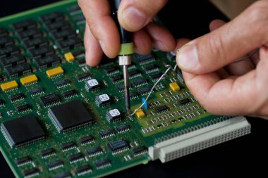 Erkek eller, elektronik bir tahtanın arka planına karşı lehim ve lehim tutarlar. Elektronik parçaların ve parçaların değiştirilmesi. Bilgisayar ekipmanları, endüstriyel ekipmanlar..