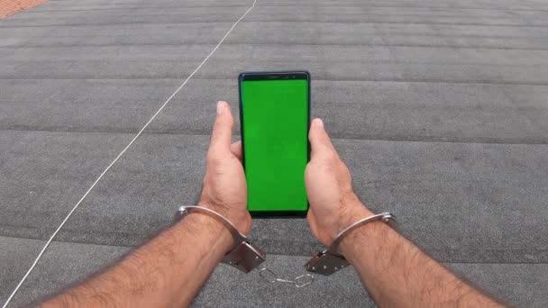 彼は手錠やチェーンを身に着けている間 垂直モードで緑の画面スマートフォンを見て道路上の若い男は 排除し 強制的な使用を取得します インターネットの閲覧 動画の視聴 ブログ — ストック動画