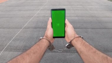 Genç adam kelepçeli ya da zincirliyken dikey modda yeşil ekranlı akıllı telefonu izleyip kurtuluyor. İnternet taraması, Videolar İzleme, Bloglar.