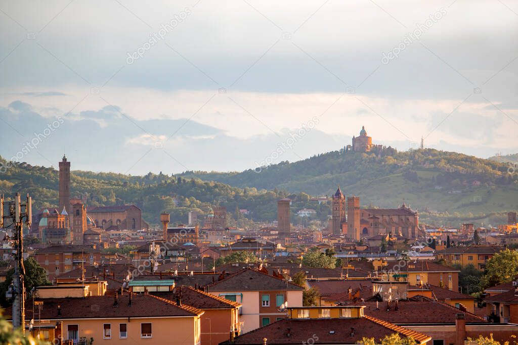 Aerial panoramic view of old Bologna city. It is saw Asinelli tower or torre, San Petronio church or basilica and San Luca church or basilica. Cloudy and sunset sky and bolognese hills in background