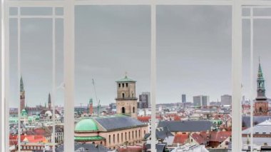 Kopenhag şehri bulutlu bir günde kuşlar uçarken beyaz bir pencereden görüldüğü gibi. Yakın planda bir kilise ve arka planda Rosenvorg şatosu kulesi görülüyor.