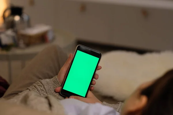 Over shoulder of one young woman holding green screen smartphone, lying on sofa  in the living room at night, Blur background