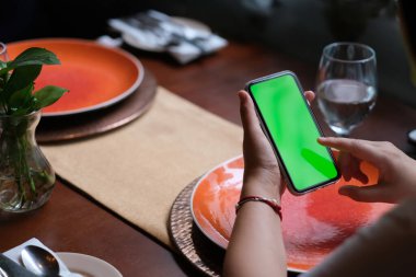 Restoranda yeşil ekran akıllı telefonu dinleyen insanların omzundan. Bulanık tabaklar ve masada bardak..