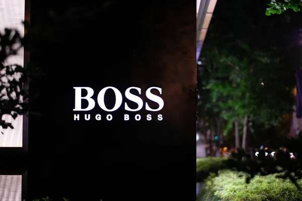 2019年7月 夜间在商店外面的墙上贴上灯火通明的Hugo Boss标志 模糊的绿色植物和街灯 一家专注于西服的德国奢侈品牌 — 图库照片