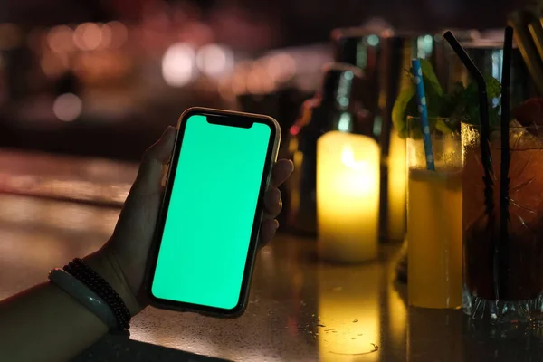 Geceleri tezgahın üzerinde yeşil ekran akıllı telefon tutarak elinizi kapatın. sönmüş mum ışığı ve kokteyl