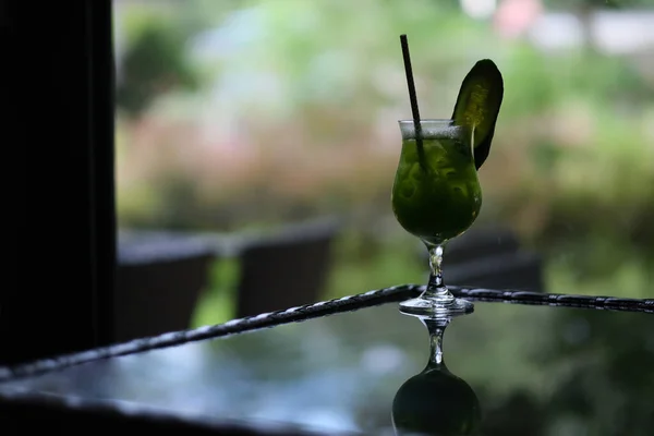 a glass of vegetable juice with slice of cucumber on table. defocused green garden plants background