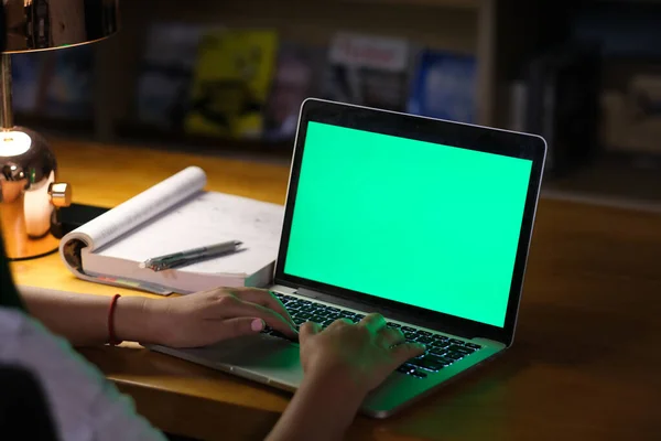 over shoulder view of people typing on green screen laptop computer with book on table under lamp at night