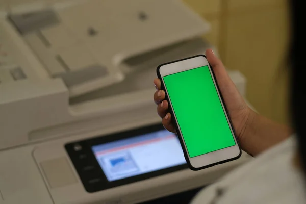 over shoulder view of young female holding green screen smart phone in office, in front of printer