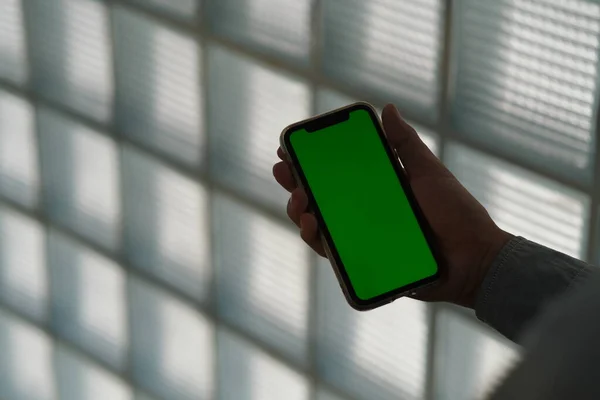 over shoulder view of business man holding green screen smart phone. Defocused modern glass wall background