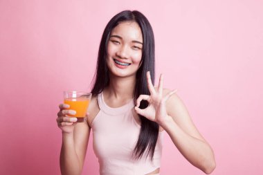 Genç Asyalı kadın portakal suyu içiyor. Pembe arka planda tamam işareti var.