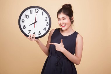 Genç Asyalı kadın bej arka planda bir saate işaret ediyor.