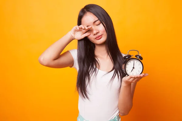 Sleepy young Asian woman with a clock in the morning on bright yellow background