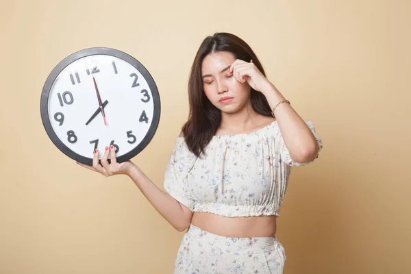 Sleepy young Asian woman with a clock in the morning on beige background