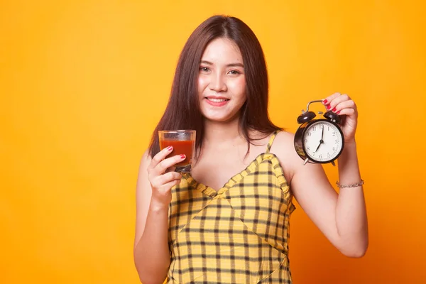 Young Asian woman with tomato juice and clock on yellow background