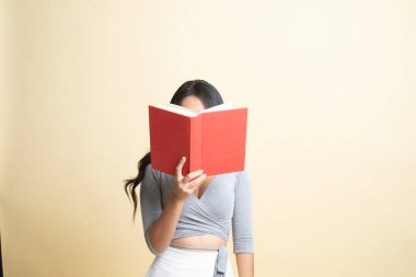 Kitabı olan genç bir Asyalı kadın bej arka planda yüzünü kapatıyor.