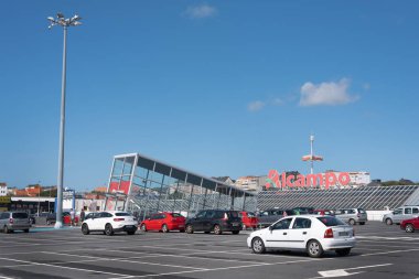 Ferrol, Galiza, İspanya - 08: 20 2020: Alcampo otoparkı, güneşli bir yaz sabahı mavi gökyüzü. Tanımadığımız iki kişi uzaklardan geçiyor. Kırmızı ve beyaz. Çok yüksek sokak lambaları