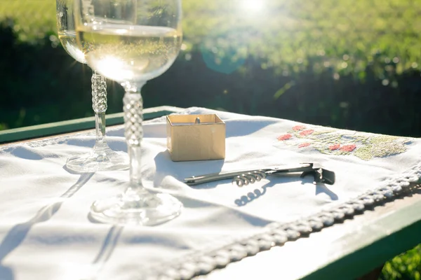 White wineglasses for toasting, golden engagement ring focused with sun rays on garden table with tablecloth and corkscrew. Copy space available. Wedding invitation background