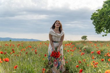 Beyaz elbiseli genç bir kadın kameraya kırmızı haşhaş çiçekleri atıyor.