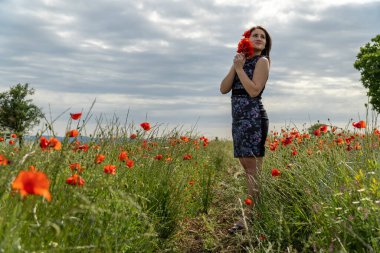 Haşhaş tarlasında mavi elbiseli genç bir kadın. Arkasında güzel bulutlar var.