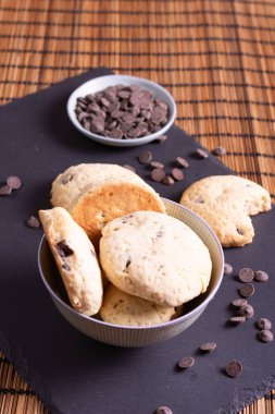 Çikolata parçalı kurabiye kasesi çikolatalı ve bir ısırık kurabiyesi bambu arka planda servis ediliyor.