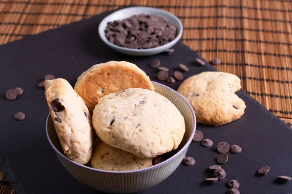 Çikolata parçalı kurabiye kasesi çikolatalı ve bir ısırık kurabiyesi bambu arka planda servis ediliyor.