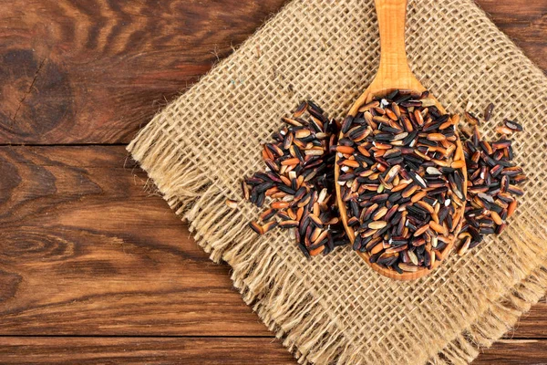 Black rice in spoon on burlap and wooden background closeup