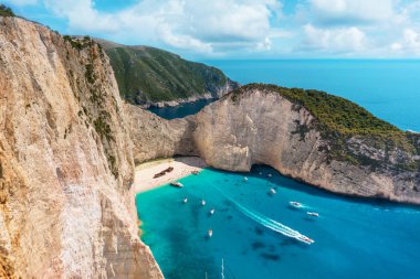 Shipwreck beach at sunny day with a huge rock on foreground, floating boats and tiny tourists on a beach. Zakynthos island, Greece. clipart