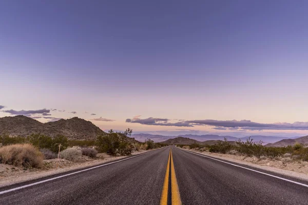 Desert road at sunset time with crazy sky and clouds, dry plants on road sides and mountains on horizon.