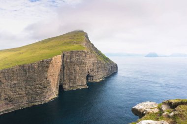 Okyanusun üzerinde yeşil dağ Kayası ve ufukta uzak adalar. Vagar, Sorvagsvatn, Faroe Adaları.