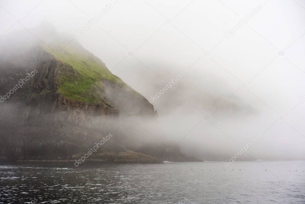 Misty landscape of Mykines island shoot from water. Fantastic green cliffs above the ocean and thick morning fog.