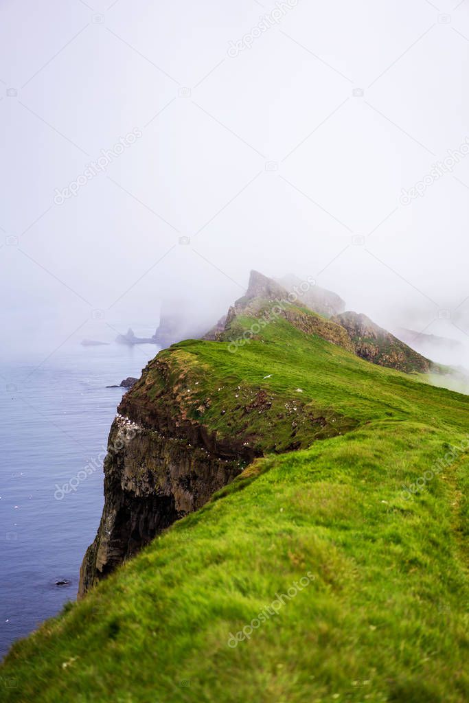Foggy landscape of green cliffs above the ocean. Mykines, Faroe Islands.