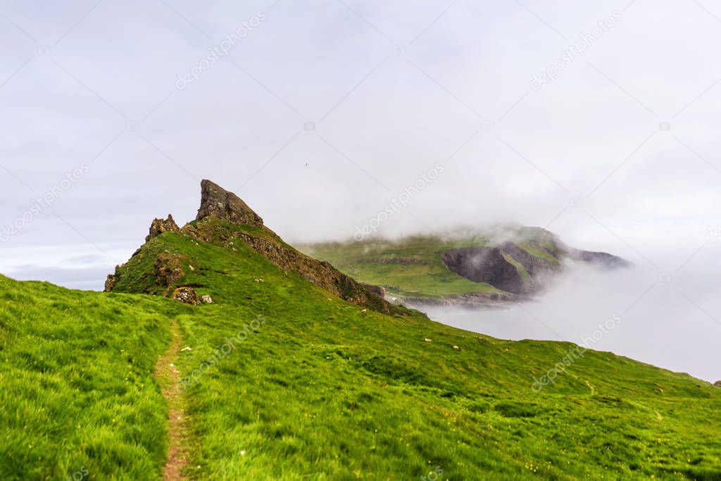 Hike path on Mykines island with green mountains and thick fog covering cliffs. Faroe Islands, Denmark.