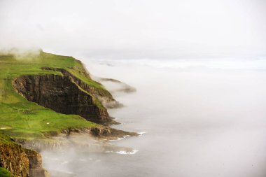 Atlas Okyanusu'nun üzerindeki Mykines adasının yeşil kayalıkları ve sudan gelen hafif sis. Faroe Adaları.