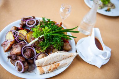 Yanında patates, çiğ soğan, maydanoz ve pide soslu et şişleriyle dolu bir tabak.