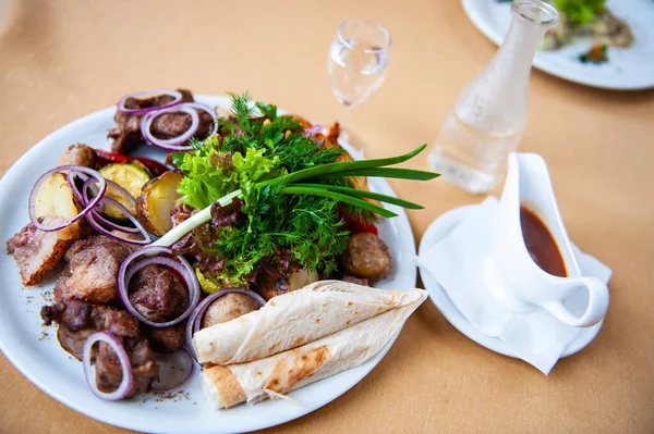 A dish full of meat skewers with a side dish of potatoes, raw onions and parsley and pita, sauce in a sauceboat