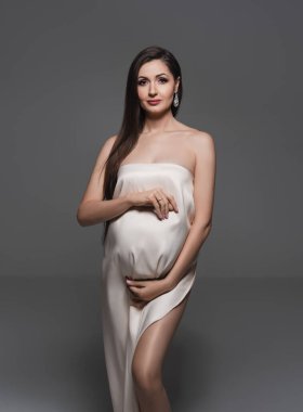 Duvağa sarılmış hamile bir kadın kollarıyla karnını tutuyor. Mutlu hamile bir kadının moda portresi. Hamile kadın ve annelik, hazırlık ve beklenti konseptine sarılmış. Yakın plan, kopyalama alanı