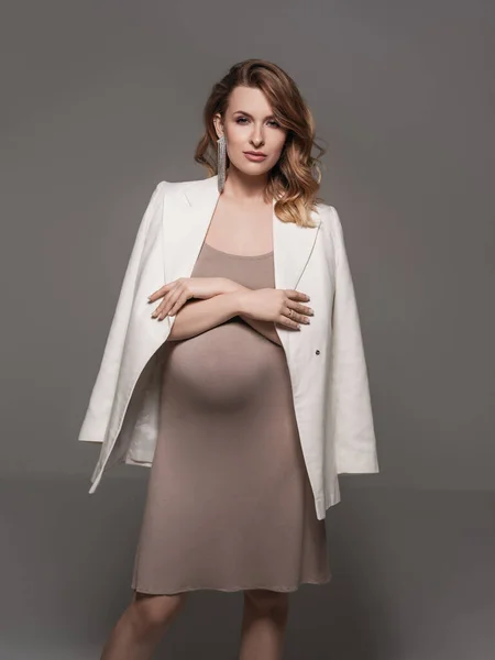 A young pregnant woman in a beautiful dress and white jacket posing in the studio.Beautiful pregnant woman touching her belly with hands on a grey background. Young mother anticipation of the baby.pregnancy fashion