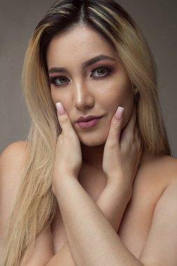 Güzel sarışın Latin model, parlak gözlü ve makyajlı, elleri yanaklarında. 