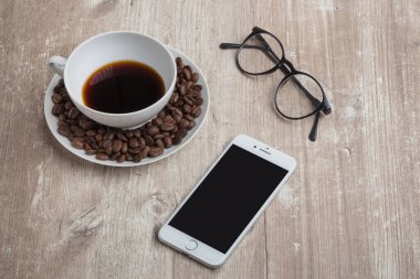 Kahve çekirdekli bir fincan, beyaz bir telefon ve eski ahşap bir masada okuma gözlüğü.