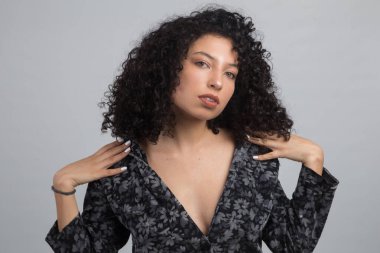 Kıvırcık saçlı, kahverengi tenli, elleri omuzlarında, siyah ve baskılı bluzu olan, gri arkaplanlı bir stüdyo fotoğrafı çeken genç Latin bir kadının yüzü.