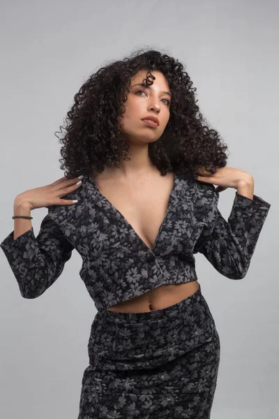 Young Latin woman with curly hair and brown skin, posing with her hands on her shoulders, wearing a black print top and skirt, studio shot with gray background