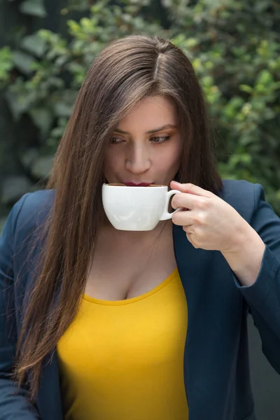 young white-skinned woman with long straight hair drinking coffee from a white cup, wearing a yellow blouse and a blue jacket, plants in the background, outdoor photo in the daytime
