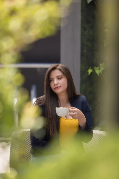 young white-skinned woman with long straight hair, holding in her hand a white cup, wearing a yellow blouse and a blue jacket, blurred bushes that are around, outdoor photo in the day