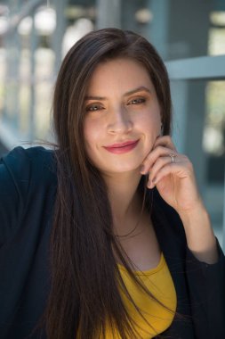 Beyaz tenli, uzun düz saçlı, açık renk gözlü ve makyajlı genç bir kadının yüzü. Yanağında bir elle gülümsüyor. Önüne bakıyor, sarı bluz ve mavi ceket giyiyor.