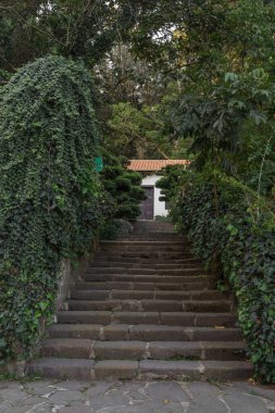 Taş basamaklı ana giriş, ağaçlar ve çalılarla çevrili, gündüz fotoğrafı.