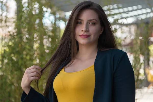 Beyaz tenli, uzun düz saçlı, açık renk gözlü ve makyajlı genç bir kadının yüzü. Bir eliyle gülümseyerek saçlarını alıyor, önüne bakıyor, sarı bluz ve mavi ceket giyiyor, arka planda bitkiler var.